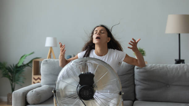 Young Caucasian woman sit on couch in living room breathe fresh air from floor fan suffering from lack of conditioner, millennial girl use ventilator struggle with home heat, summer hot weather