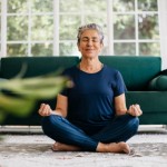 Senior woman meditating in lotus position at home, sitting on the floor in fitness clothing. Mature woman doing a breathing workout to achieve relaxation, peace and mindfulness.